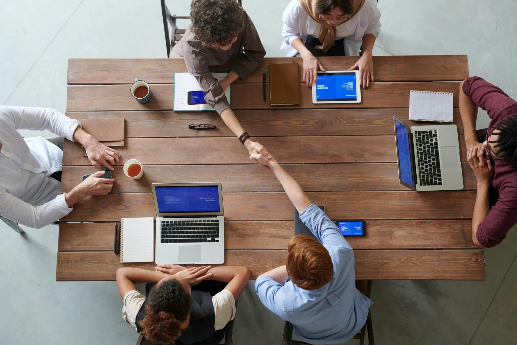 A picture of board meeting with members shaking hands, using laptops and drinking coffee.