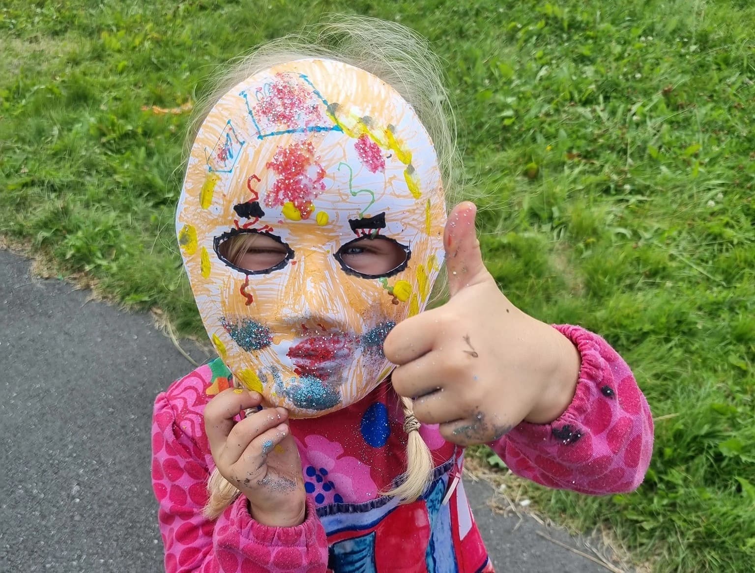 A young girl winking behind a colourful mask she’s made during an activity session with her thumb up.